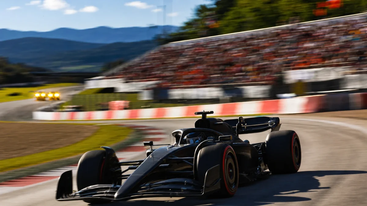 Formel 1-bil på Red Bull Ring i eftermiddagsljus med gula flaggljus tända vid en kurva i bakgrunden.